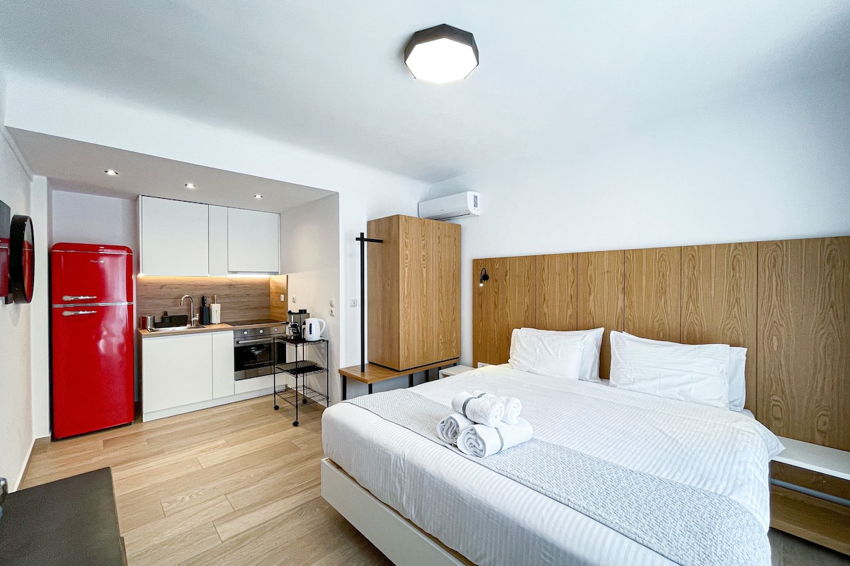 Une chambre d’hôtel moderne avec un lit, une tête de lit en bois, une kitchenette dotée d’un réfrigérateur rouge et un plancher en bois.