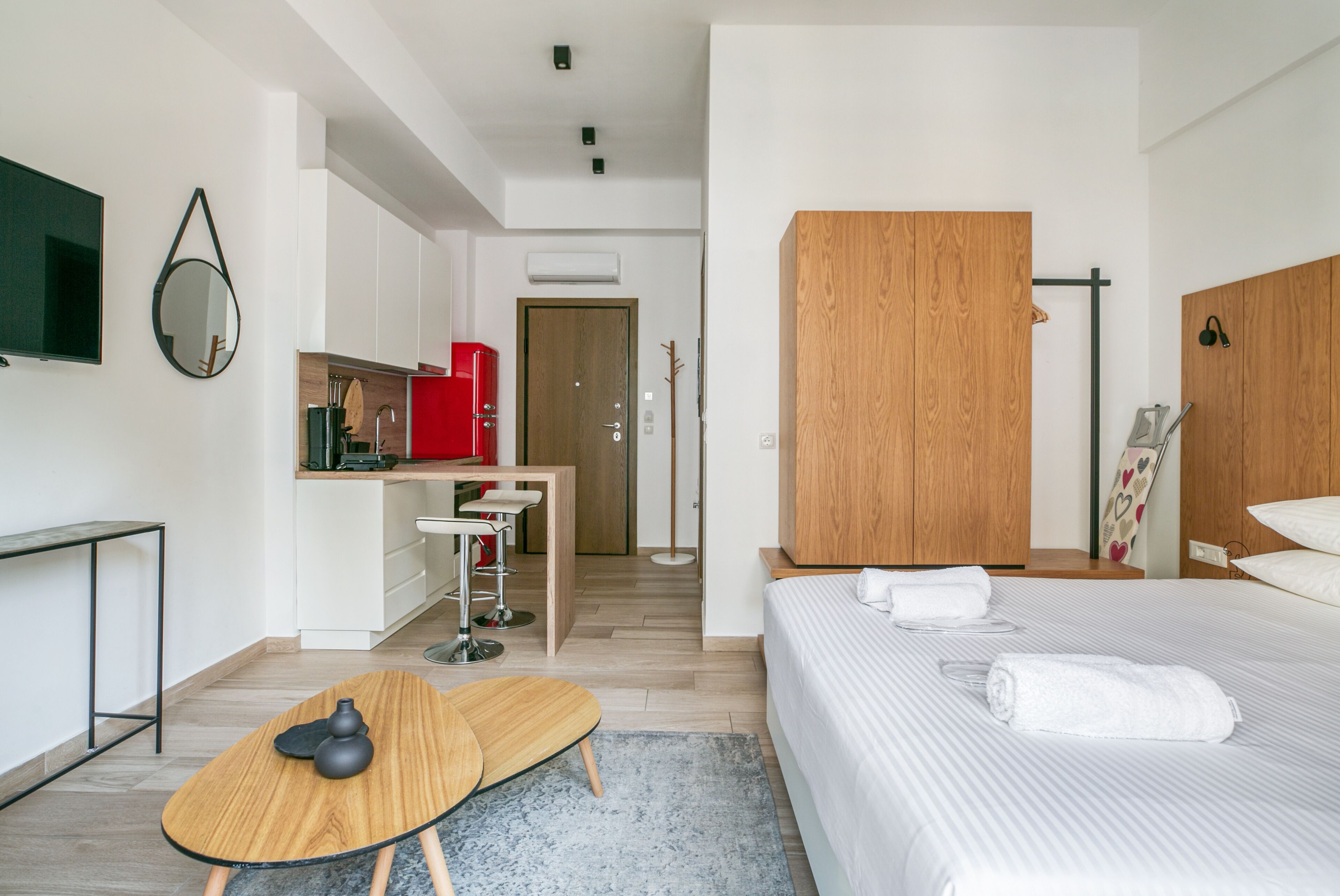 A modern hotel room with a wooden wardrobe, a bed with white linens, a small desk with a red appliance, and a mirror on the wall.
