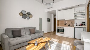 Un salon moderne avec un canapé gris, une table basse en bois et un coin cuisine doté de meubles blancs et d’un four encastré.