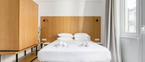 A modern hotel room with a wooden headboard, a large bed with white linens, a wooden wardrobe, and a view of the city through the window.