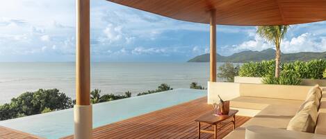 A wooden deck with a view of the sea and mountains, a lounge area with a table and cushions, and a clear sky.