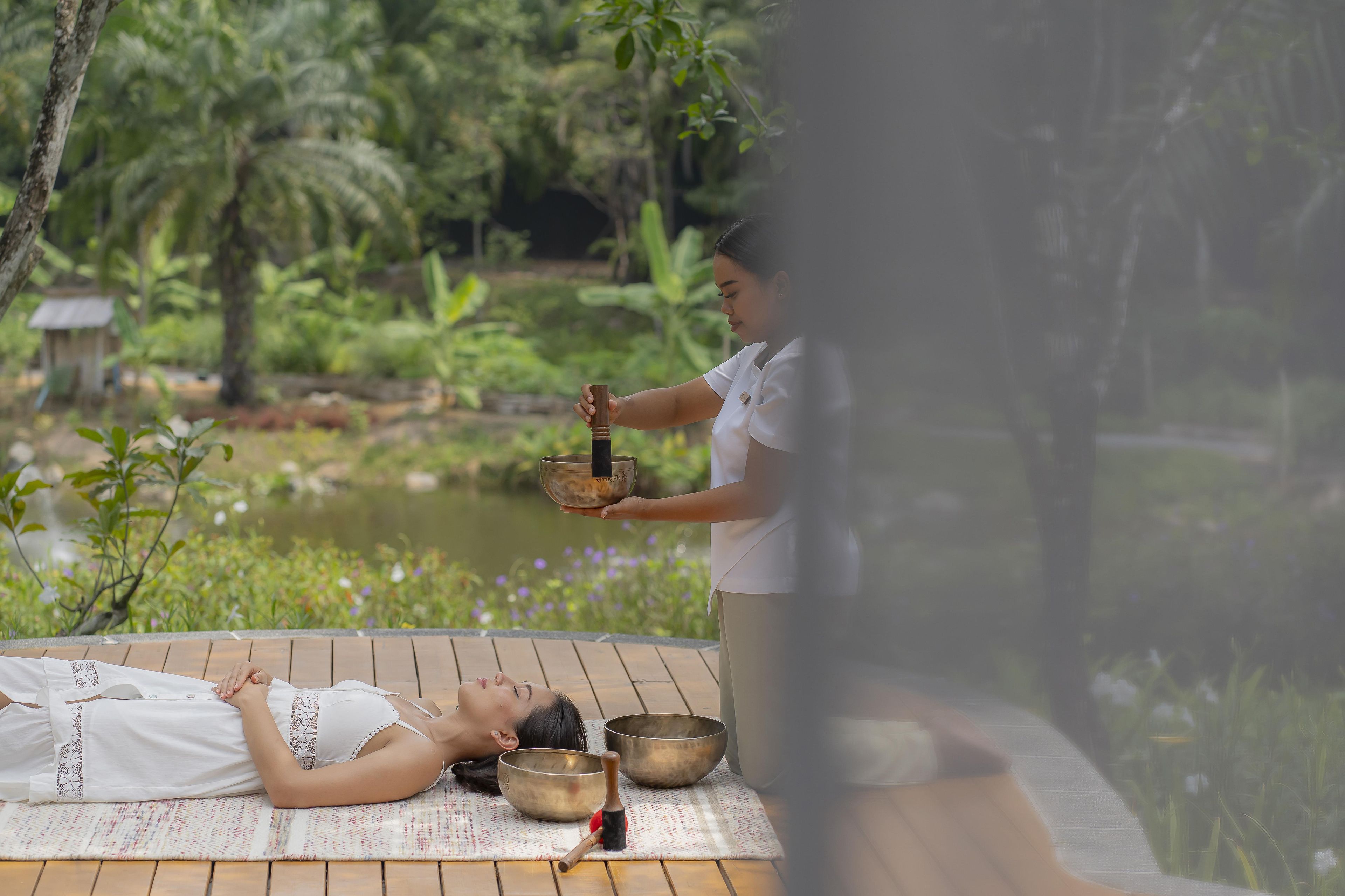 A person lying on a mat outdoors with a bowl and mallet, another person standing nearby, and a serene natural background.