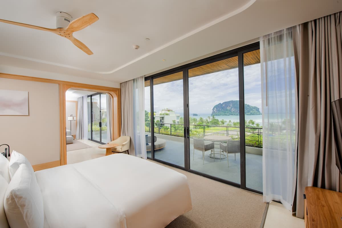 A hotel room with a large bed, a ceiling fan, a view of a balcony with outdoor furniture, and a large window.