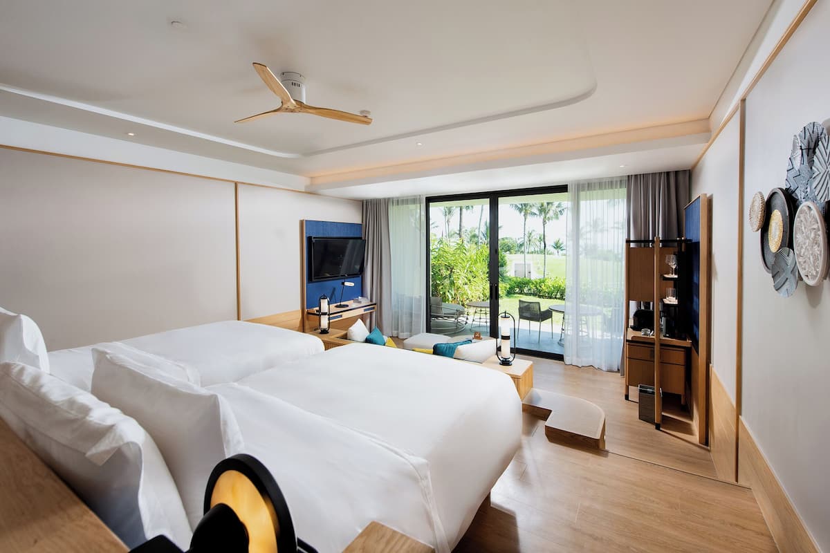 A modern hotel room with a large bed, a TV, a desk, and a balcony with a view of greenery.