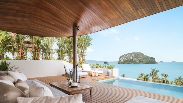 A wooden deck with a pool, white cushions, and a view of the sea and a small island.