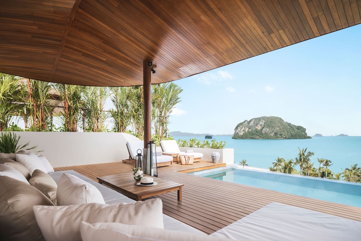 A wooden deck with a pool, white cushions, and a view of the sea and a small island.