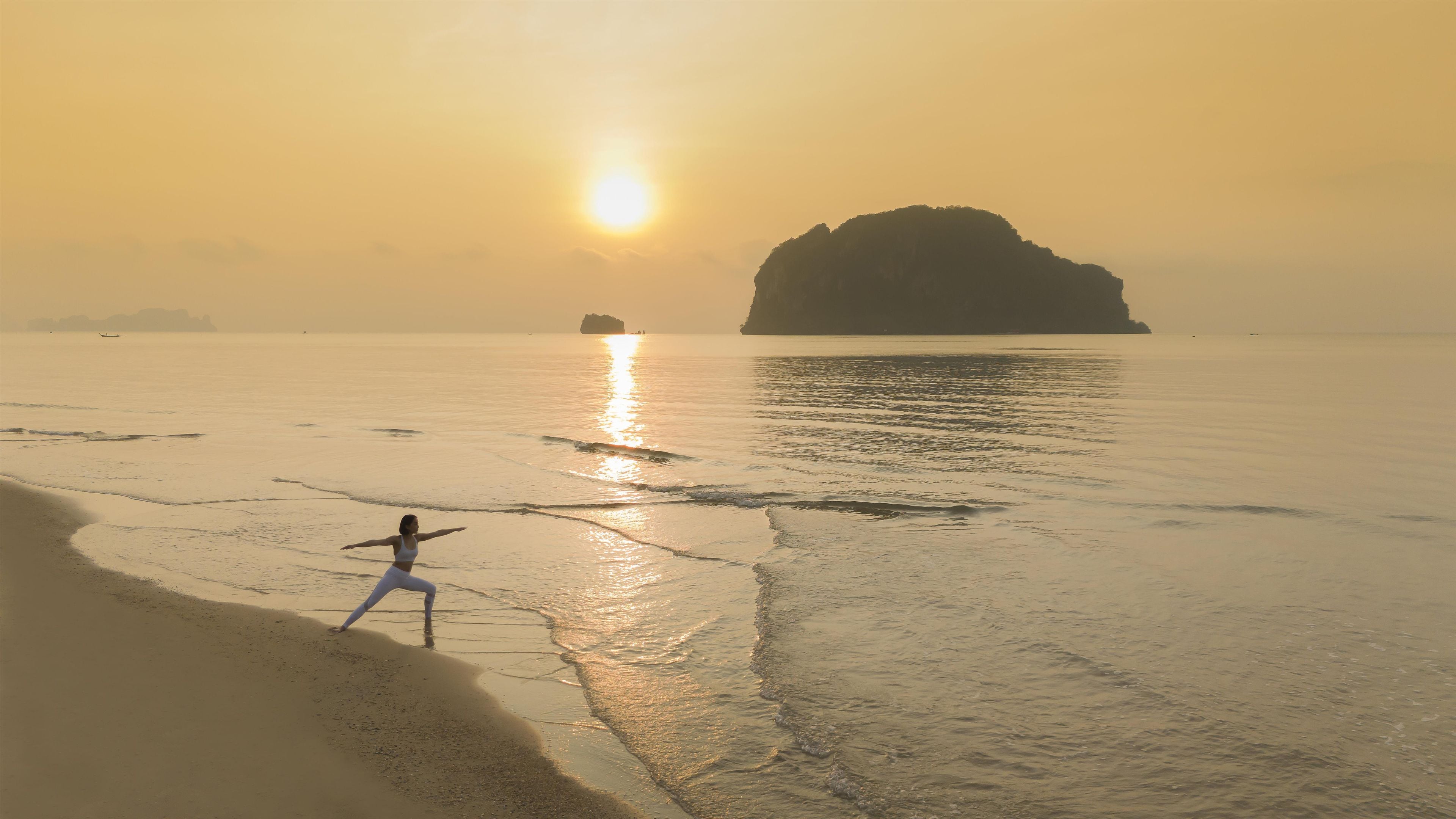 A beach scene with a person standing on the shore, waves gently lapping at the sand, and a large rock formation in the distance.