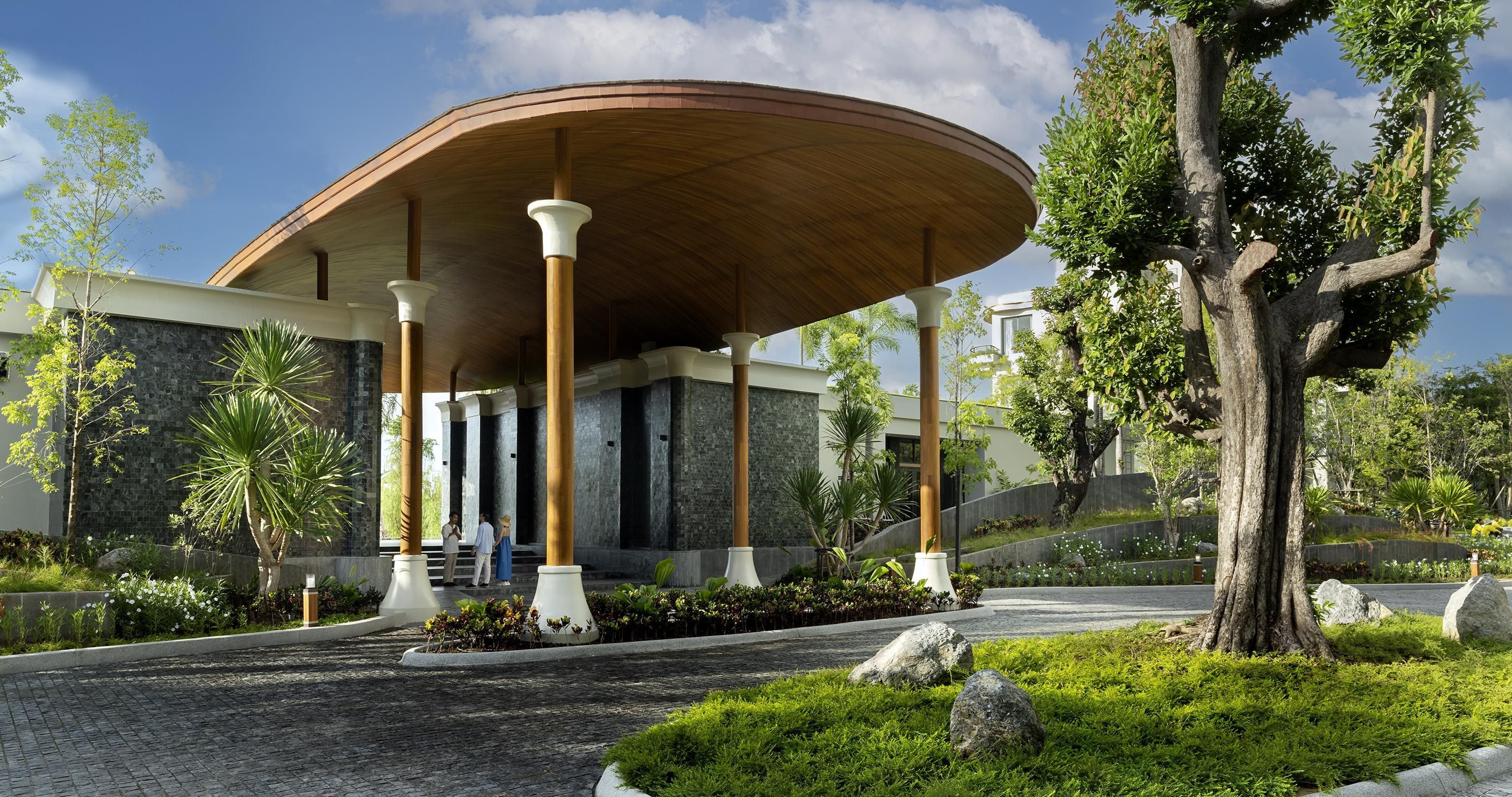 Un bâtiment moderne doté d’un toit incurvé, de murs en pierre et d’un jardin aménagé avec des rochers et de la verdure.
