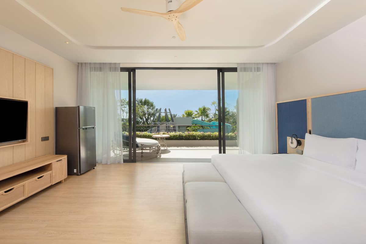 A modern hotel room with a large bed, a flat-screen TV, a refrigerator, and a balcony with a view of the outdoors.
