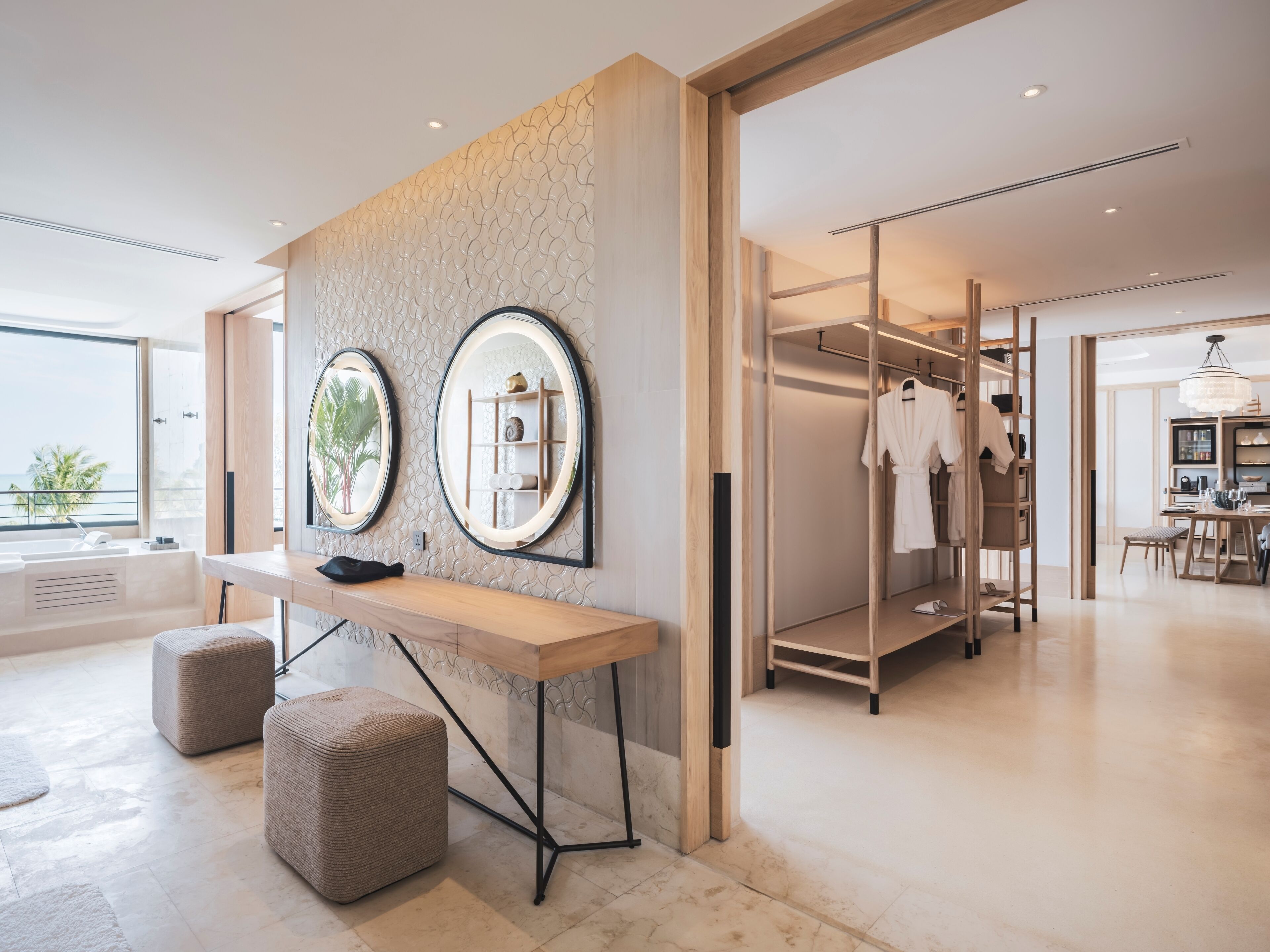 A modern hotel room with a large window, a vanity with a mirror, a robe rack, and a dining area.