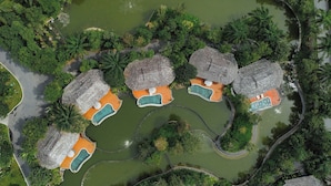 An aerial view of a resort with multiple thatched-roof cottages, each with an attached pool, surrounded by lush greenery and water features.