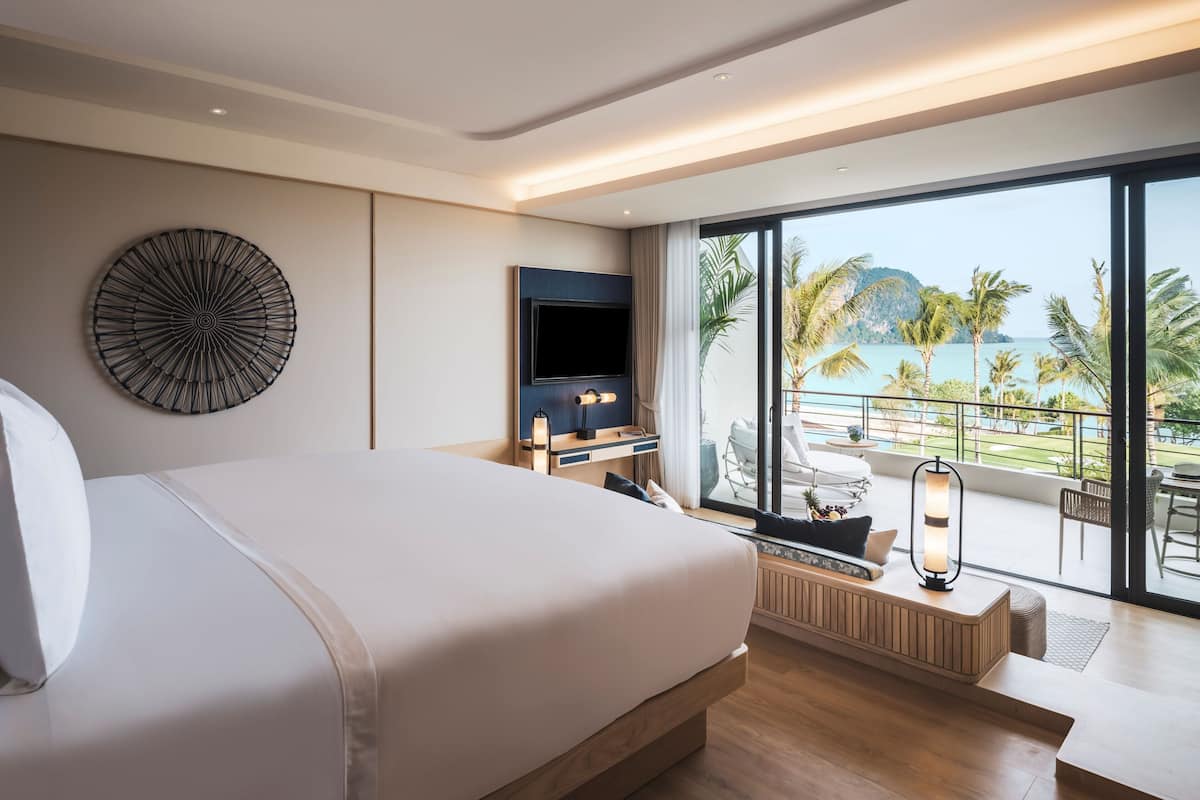 A modern hotel room with a large bed, a flat-screen TV, a wall art piece, and a balcony with a view of the sea and palm trees.