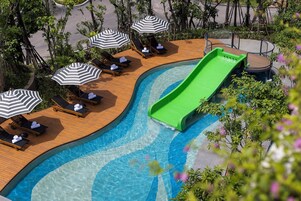 Un área de piscina con sillas de descanso y sombrillas, un tobogán verde y una cubierta de madera.