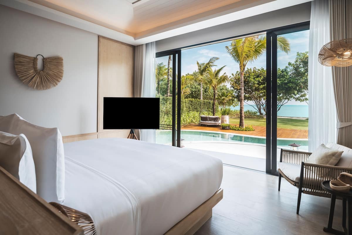 A modern bedroom with a large bed, a TV, and a view of a pool and tropical landscape.