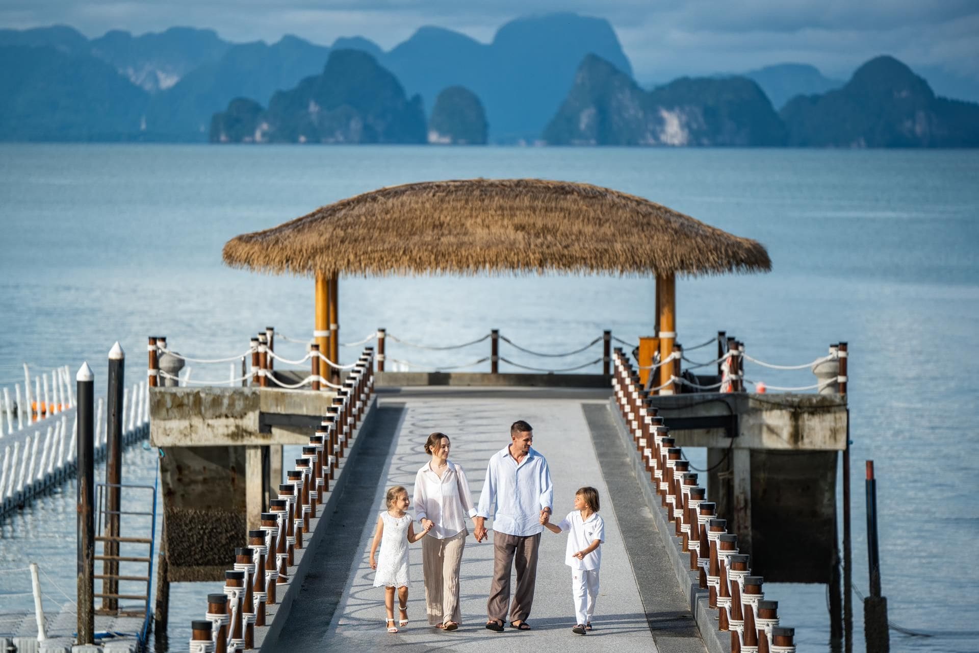 A pier with a thatched roof structure, a concrete walkway, and a view of the water and distant mountains.