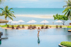 Zona de piscina con sillones de descanso y sombrillas, una mujer en traje de baño y vista a la playa.