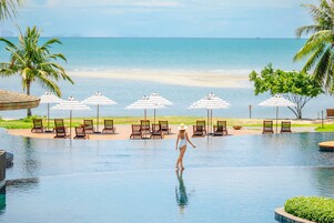 Zona de piscina con sillones de descanso y sombrillas, una mujer en traje de baño y vista a la playa.