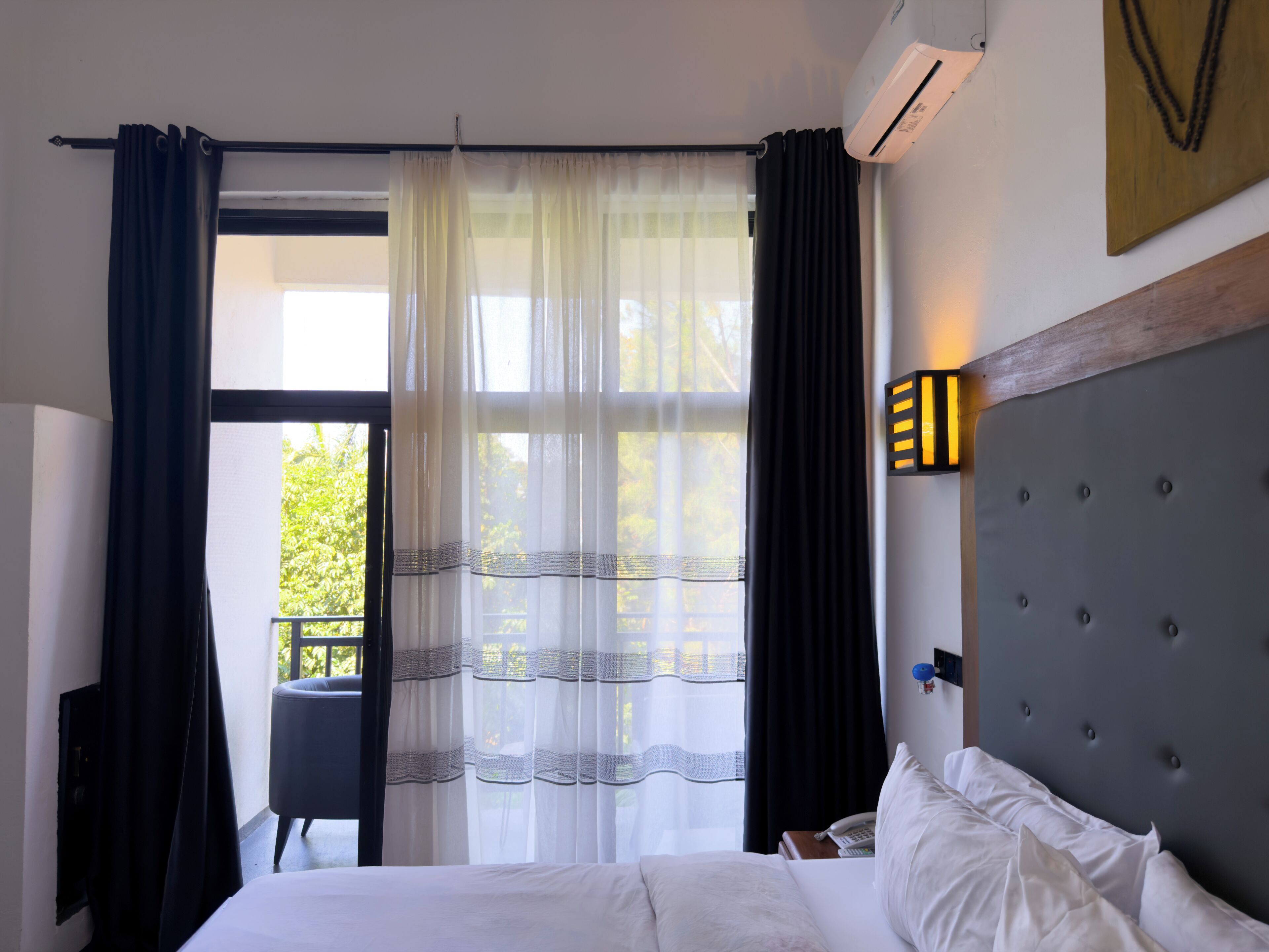 Una habitación de hotel con una cama, una silla, un ventanal con cortinas y una lámpara en la pared.