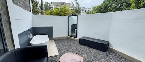Un patio moderno al aire libre con un sofá de cuero negro, una mesa redonda de madera y un lavamanos blanco.
