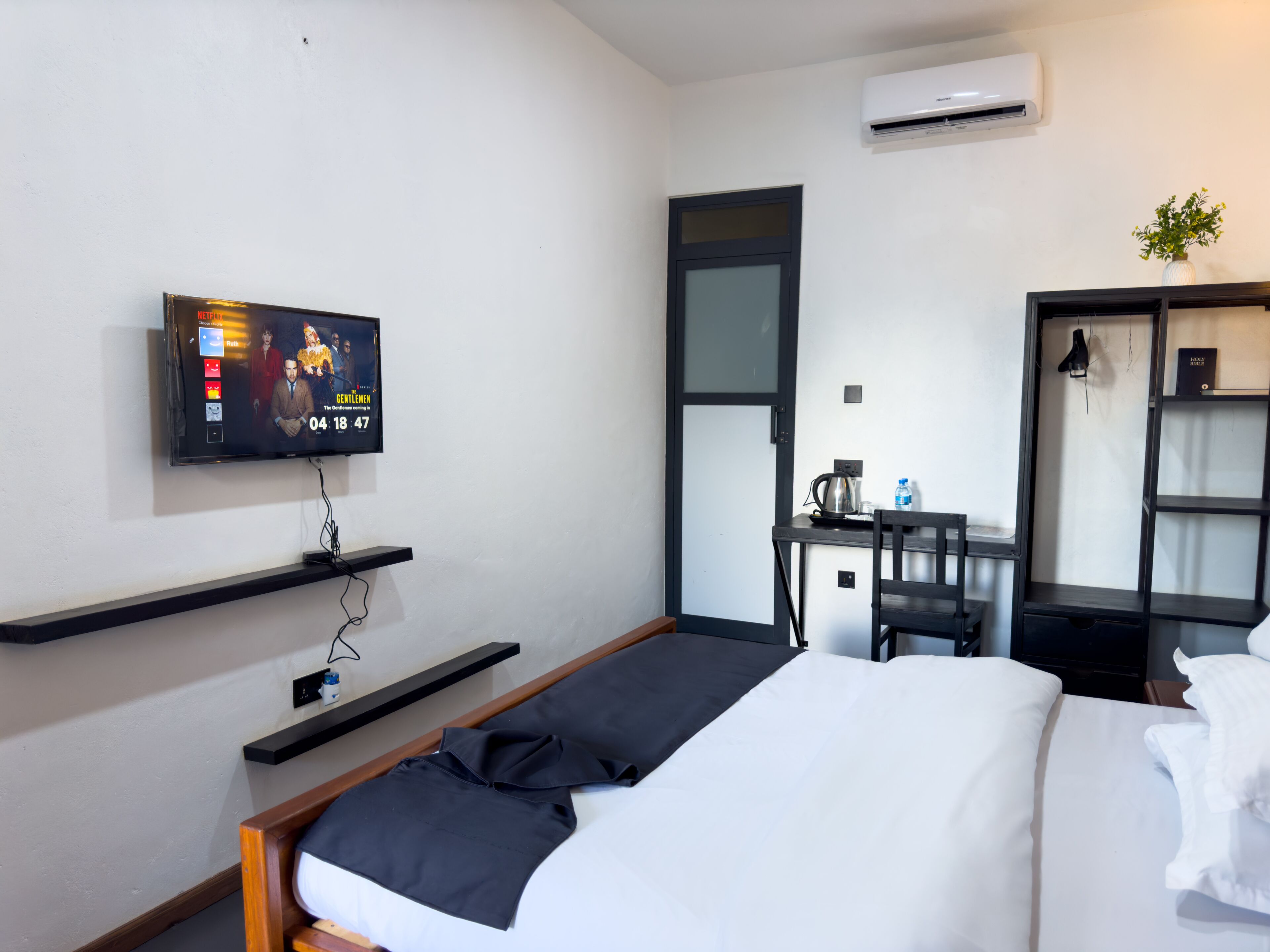 A hotel room with a bed, a TV mounted on the wall, a desk with a chair, and a small plant.