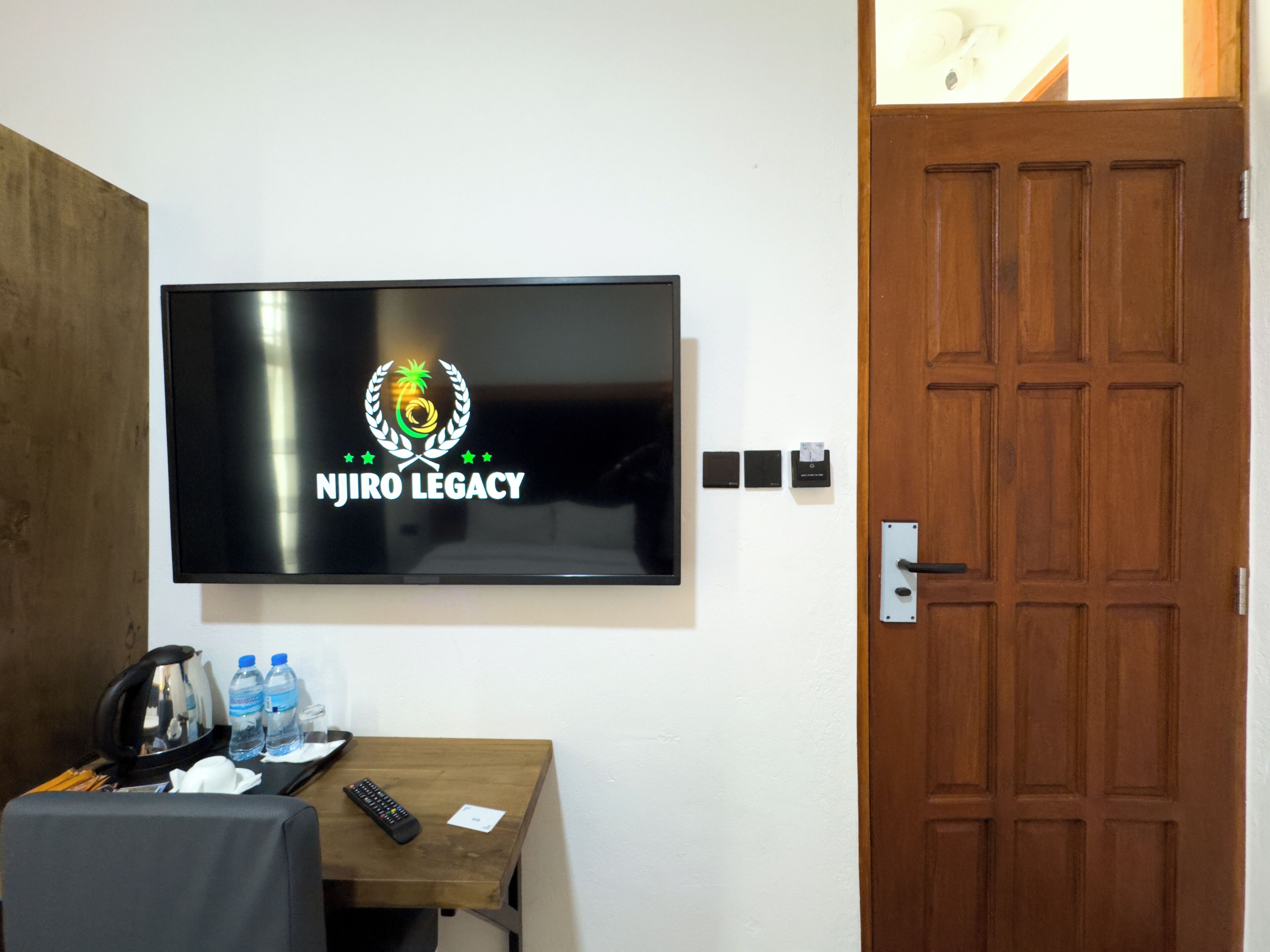 Un televisor montado en la pared mostrando el logotipo 'NJIRO LEGACY', una puerta de madera y una pequeña mesa con un control remoto y botellas de agua.