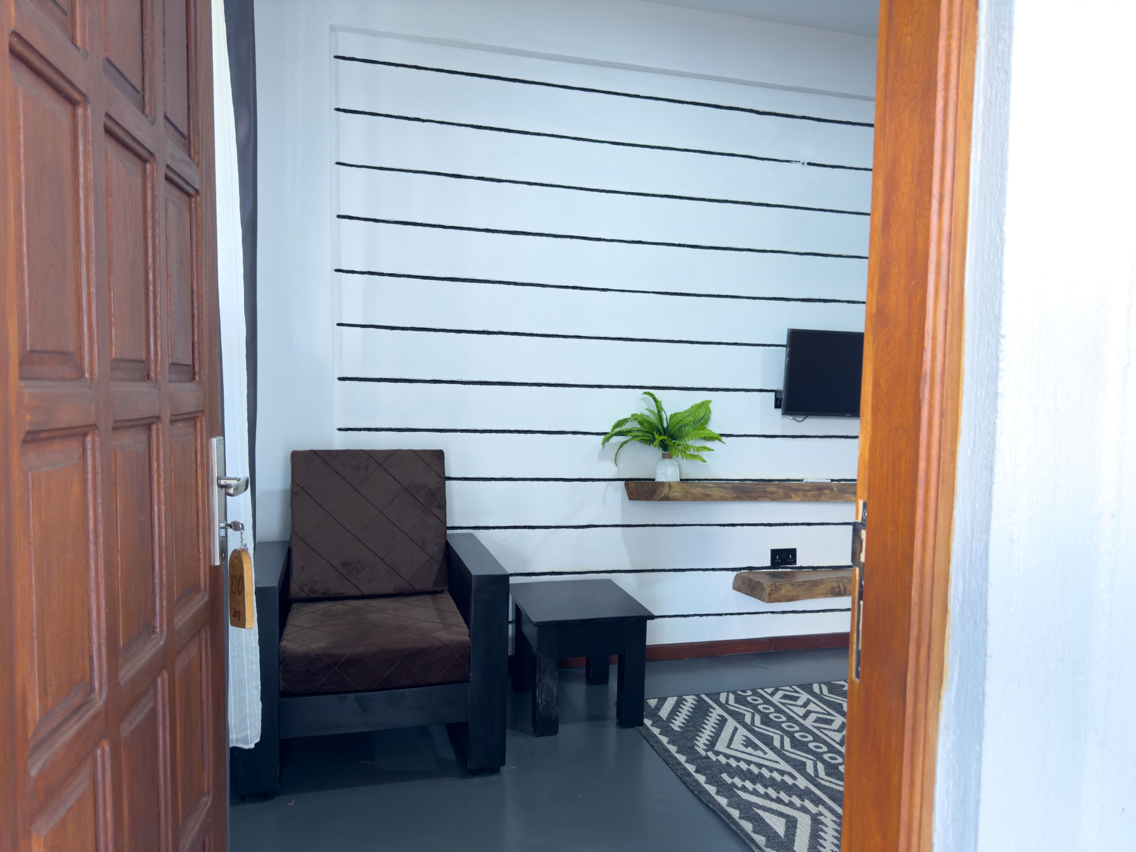 A room with a wooden door, a brown upholstered chair, a small table, a patterned rug, a wall-mounted TV, and a shelf with a plant.