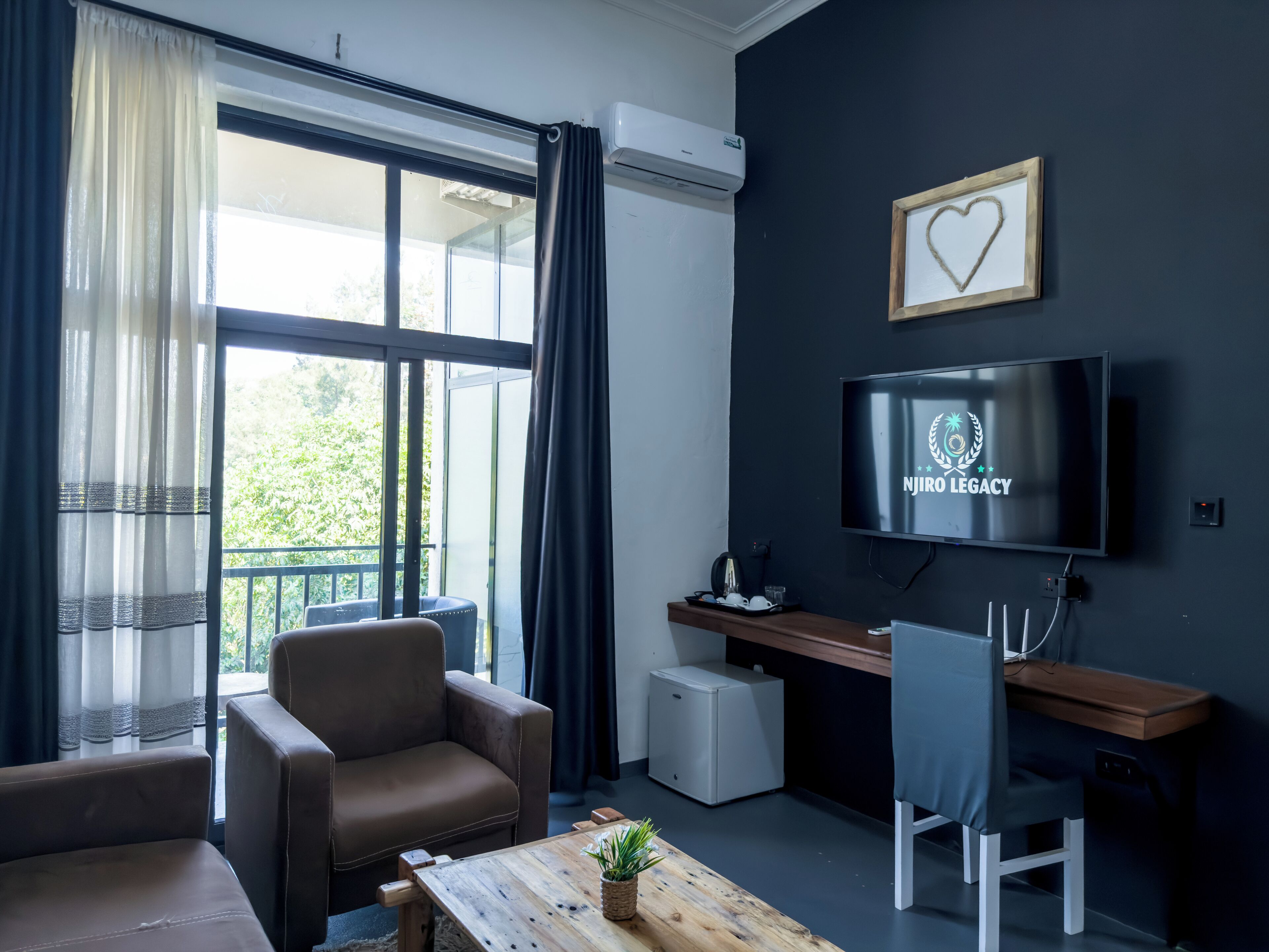 A modern living room with a sofa, a coffee table, a TV displaying "NJIRO LEGACY", and a balcony with curtains.