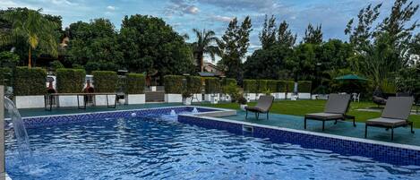 Una piscina con bordes de azulejos, rodeada de sillones de descanso y un jardín cuidado.
