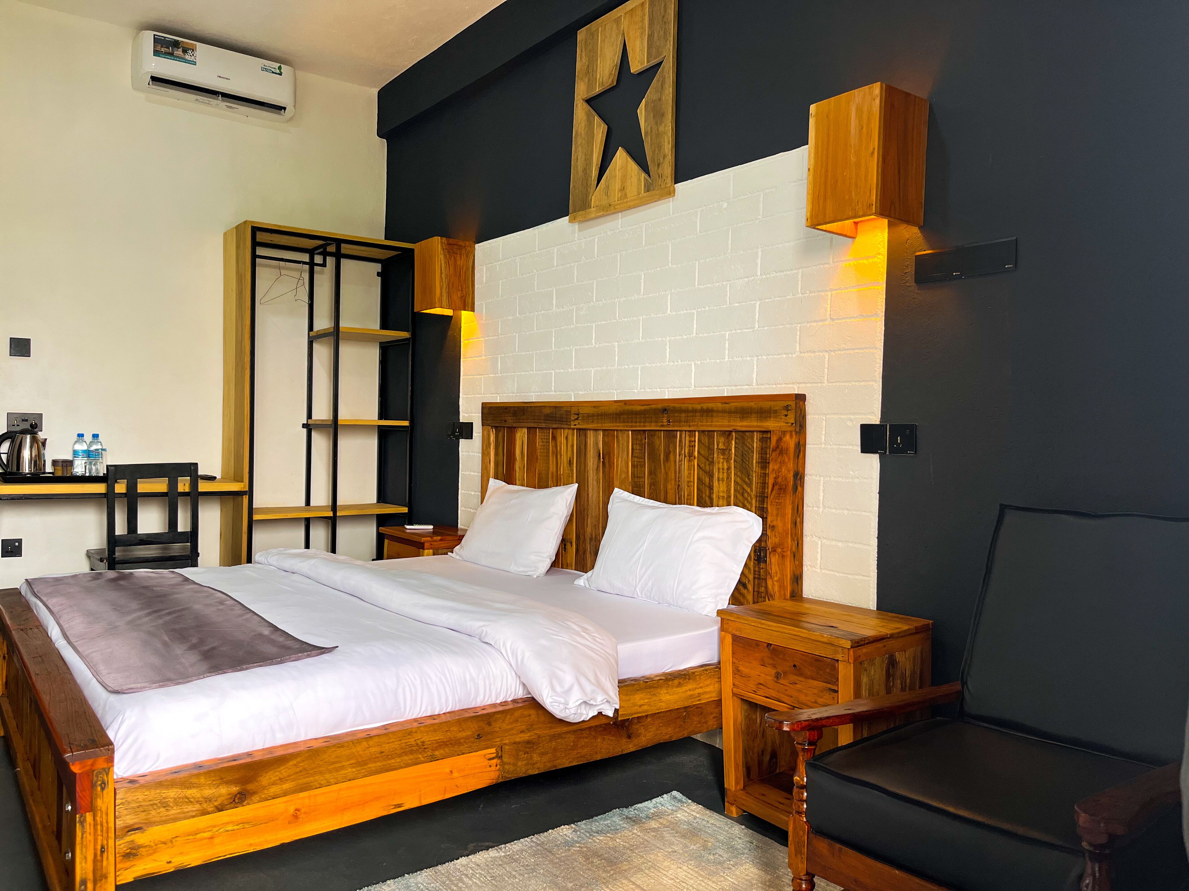 Camera d'albergo con un letto di legno, biancheria bianca, un muro in mattoni, una decorazione a stella e una scrivania con bollitore e bottiglie d'acqua.