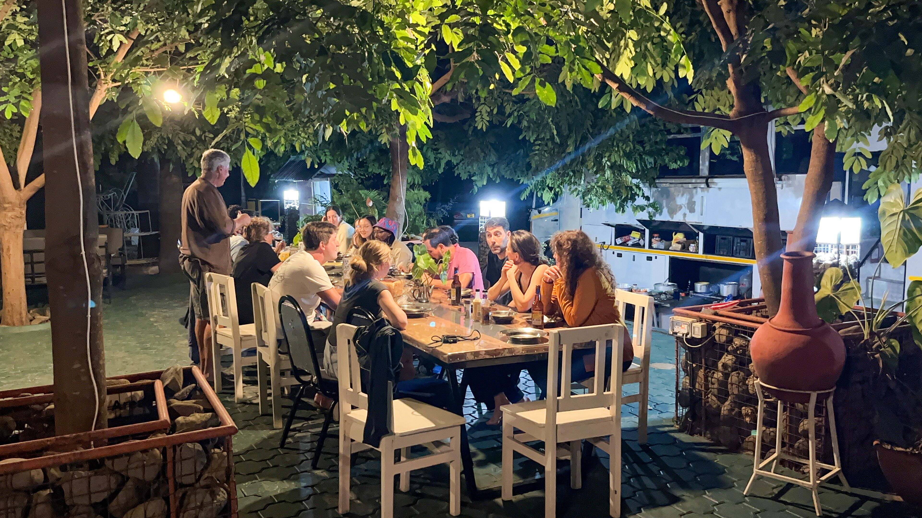 An outdoor dining area with a long table surrounded by chairs, under a tree canopy.