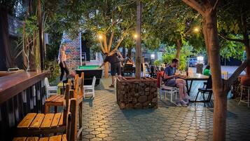 An outdoor seating area with tables and chairs, surrounded by trees and a stone wall.