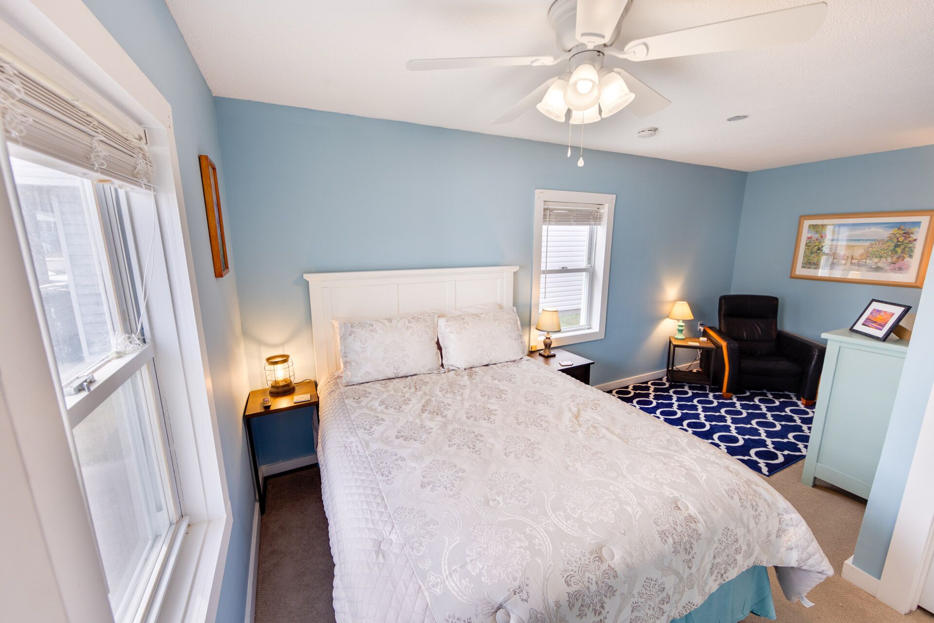Une chambre à coucher comprenant un lit, un ventilateur de plafond, une table de chevet avec une lampe, une chaise, une fenêtre avec des stores et un tableau encadré accroché au mur.
