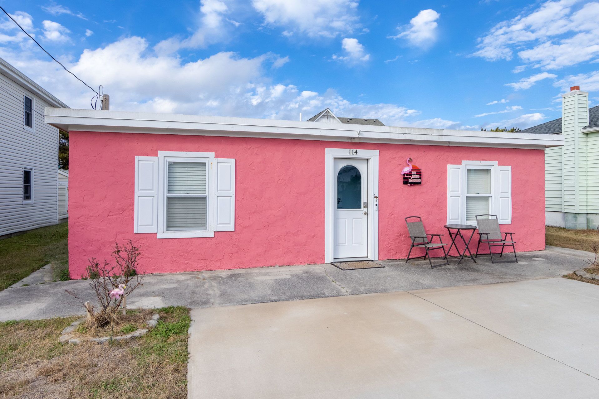 A pink house with a white door, two windows with shutters, and a small porch with two chairs.