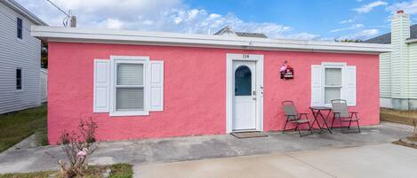 A pink house with a white door, two windows with shutters, and a small porch with two chairs.
