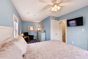 Quarto com cama, escrivaninha, cadeira, cômoda, ventilador de teto e televisão fixada na parede.