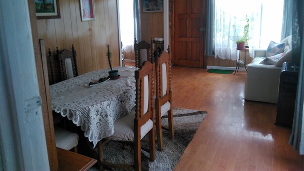 Una habitación con piso de madera, una mesa de comedor con mantel de encaje, una silla de madera, un sofá y una puerta que da a otra habitación.