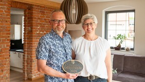 A man and a woman holding a plaque in a room with exposed brick walls, a modern pendant light, and a view of the outside through a window.