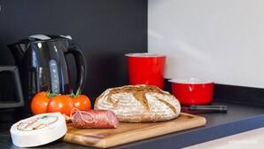 A kitchen counter with a loaf of bread, tomatoes, a cheese, a red cup, and a black kettle.