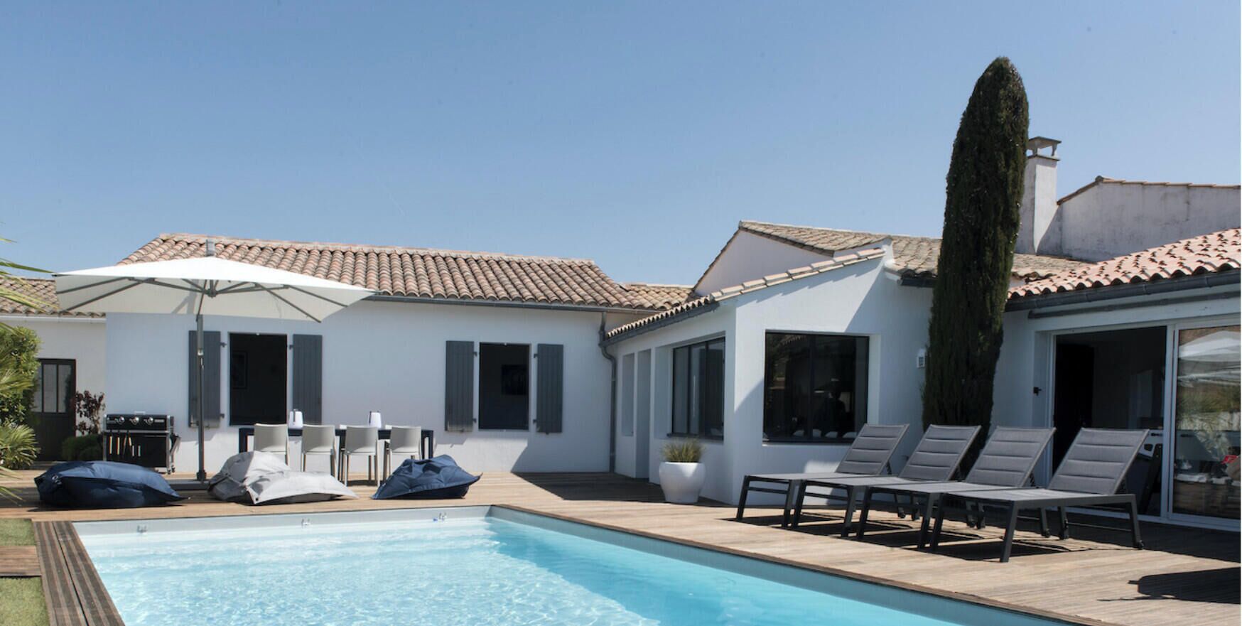 A swimming pool with lounge chairs, a barbecue area, and a white building with a tiled roof.