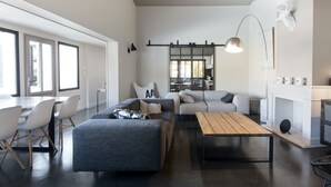 A modern living room with a sofa, ottoman, and coffee table, a dining area with chairs, and a kitchen in the background. - Comfortable villa for 10 with a pool (La Flotte)