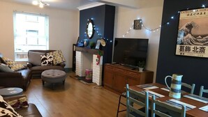 A living room with a sofa, ottoman, and a dining table with chairs. There is a fireplace with a clock and a TV on a stand. - Spacious 3 bedroom house close to the beach (Lowestoft)
