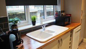 A kitchen with a white sink, a window with a view of buildings, a potted plant, and a microwave. - Spacious 3 bedroom house close to the beach (Lowestoft)