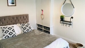 A bedroom with a tufted headboard, a bed with patterned pillows, a wardrobe, a round mirror, and a small plant on a shelf. - Spacious 3 bedroom house close to the beach (Lowestoft)