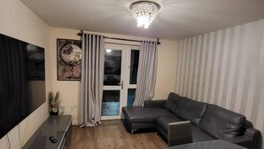 A modern living room with a black leather sofa, a flat-screen TV, a dining table with chairs, and a chandelier. - Lovely 4 Bedroom Home With Private Parking (Grays)