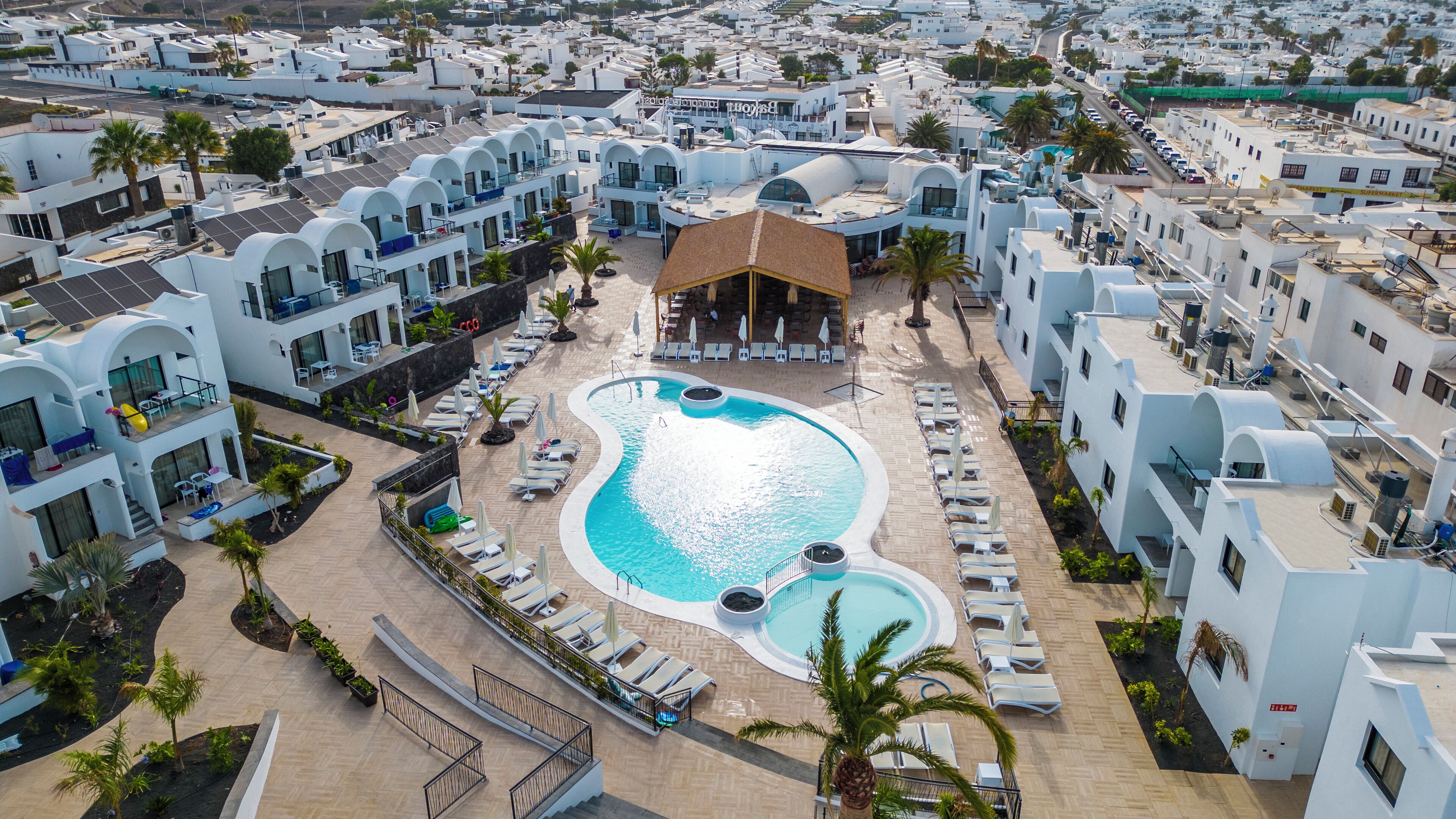 Ein Luftbild eines Resorts mit einem zentralen Swimmingpool, mehreren weißen Gebäuden und Palmen.