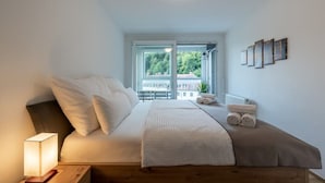 Ein Schlafzimmer mit einem Bett, Kissen, einem Nachttisch mit Lampe, einem Fenster mit Blick und einer Wand mit gerahmten Bildern.