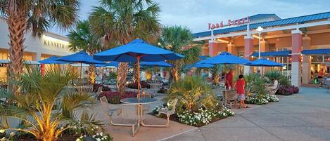 An outdoor dining area with umbrellas and chairs, a Kenneth Cole store in the background, and a pedestrian walkway.
