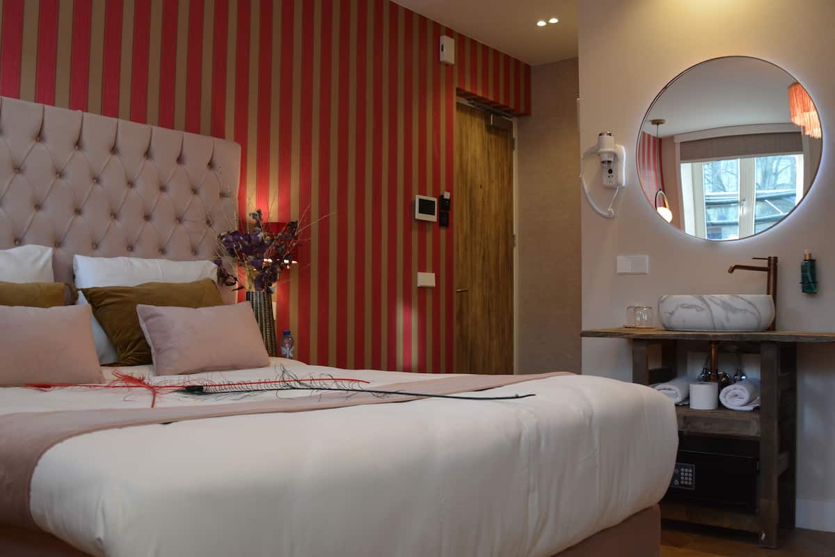 A hotel room with a tufted headboard, a large round mirror, a vanity with toiletries, and a window with a view.