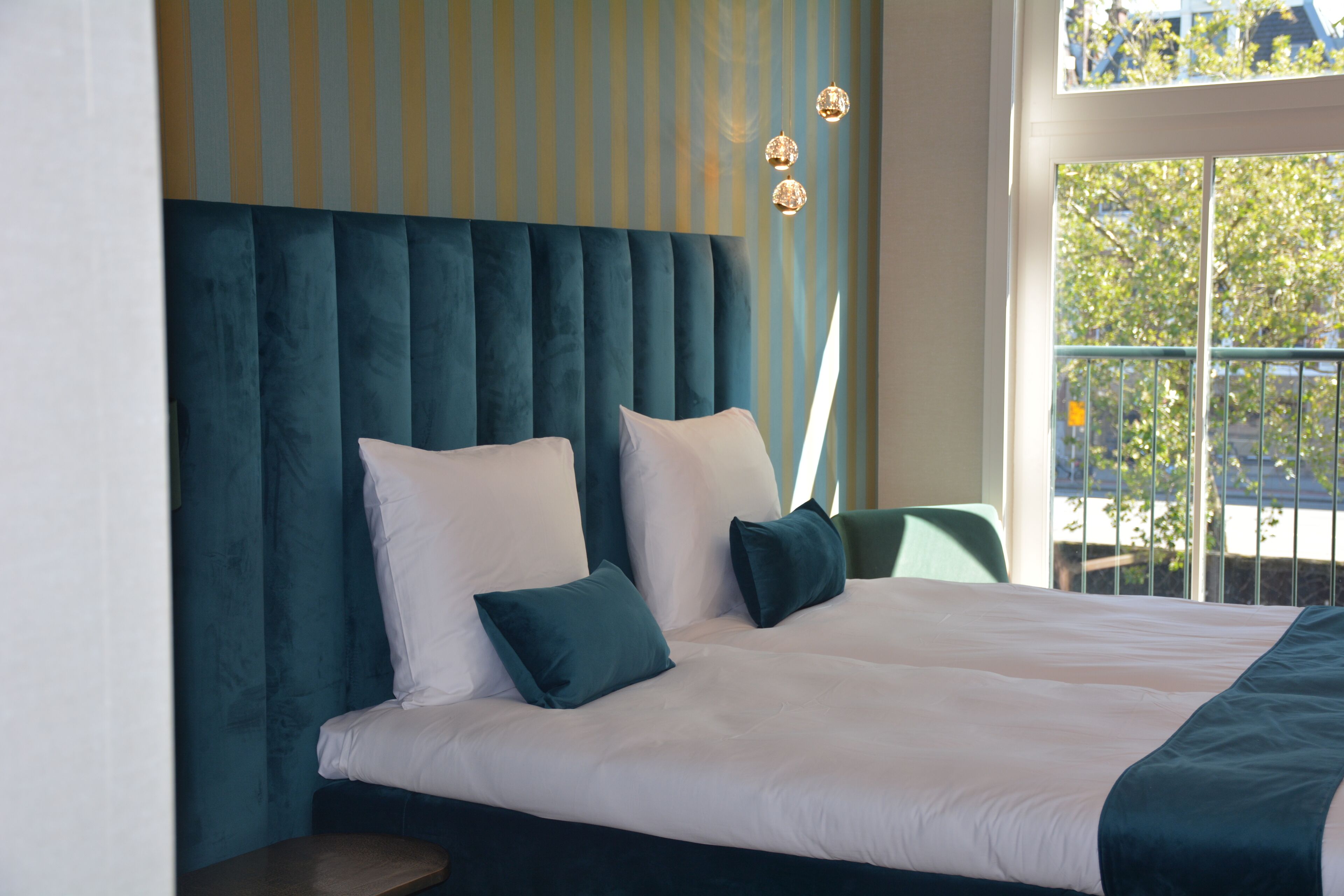A modern hotel room with a large bed, a headboard with vertical lines, and a view of the outdoors through a window.