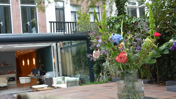 A patio with a glass vase of flowers, a seating area with a table and chairs, and a building with large windows and a balcony in the background. - If boutique hotel (Amsterdam)